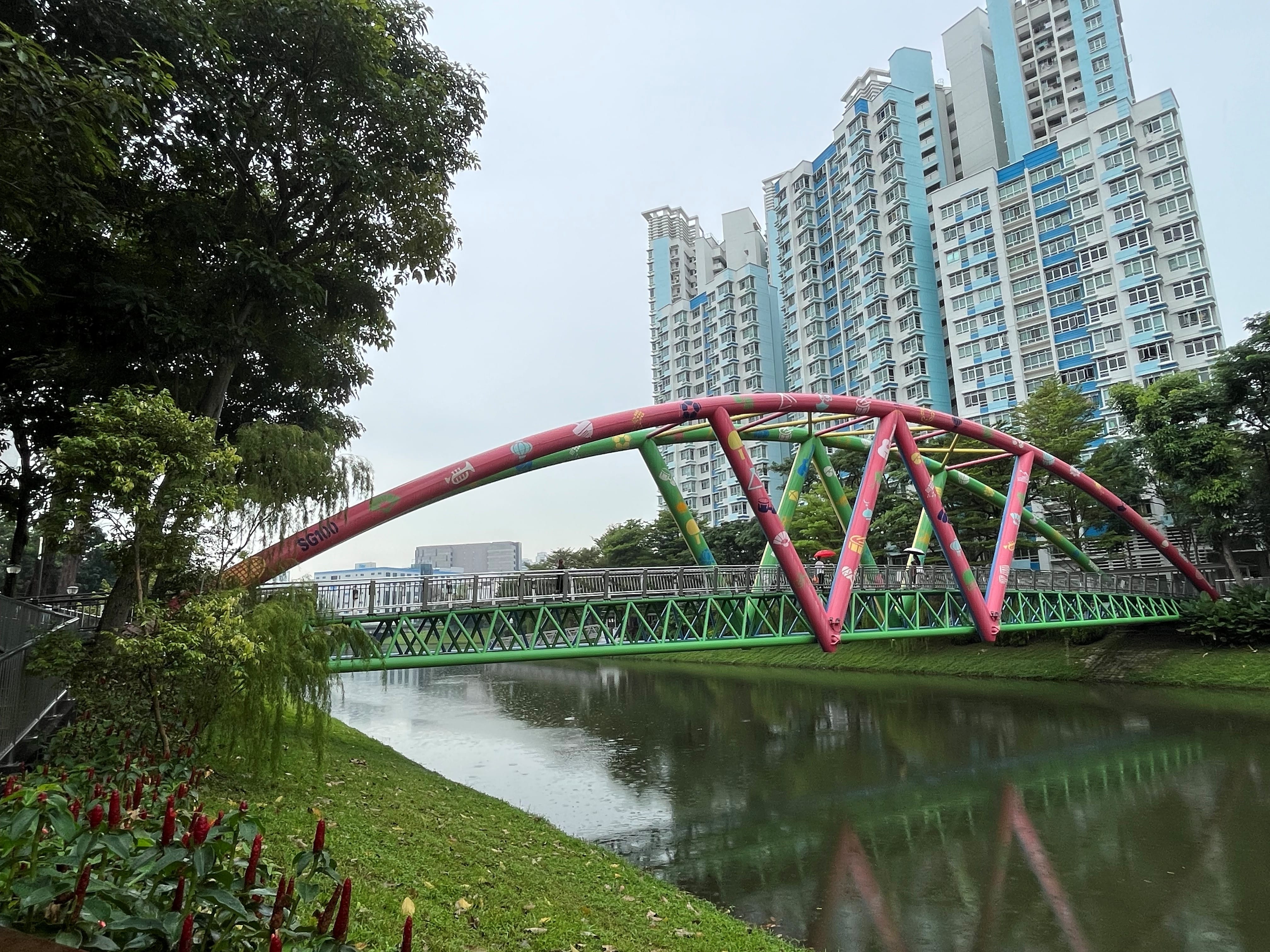 A colorful bridge spans over a canal with tall residential buildings in the background and greenery on the sides.